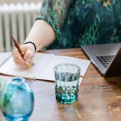 Schreibtisch mit Laptop, einem grünen Wasserglas und einer türkisfarbenen Glasvase, Maria Squarra schreibend im Bullet Journal, daneben Laptop
