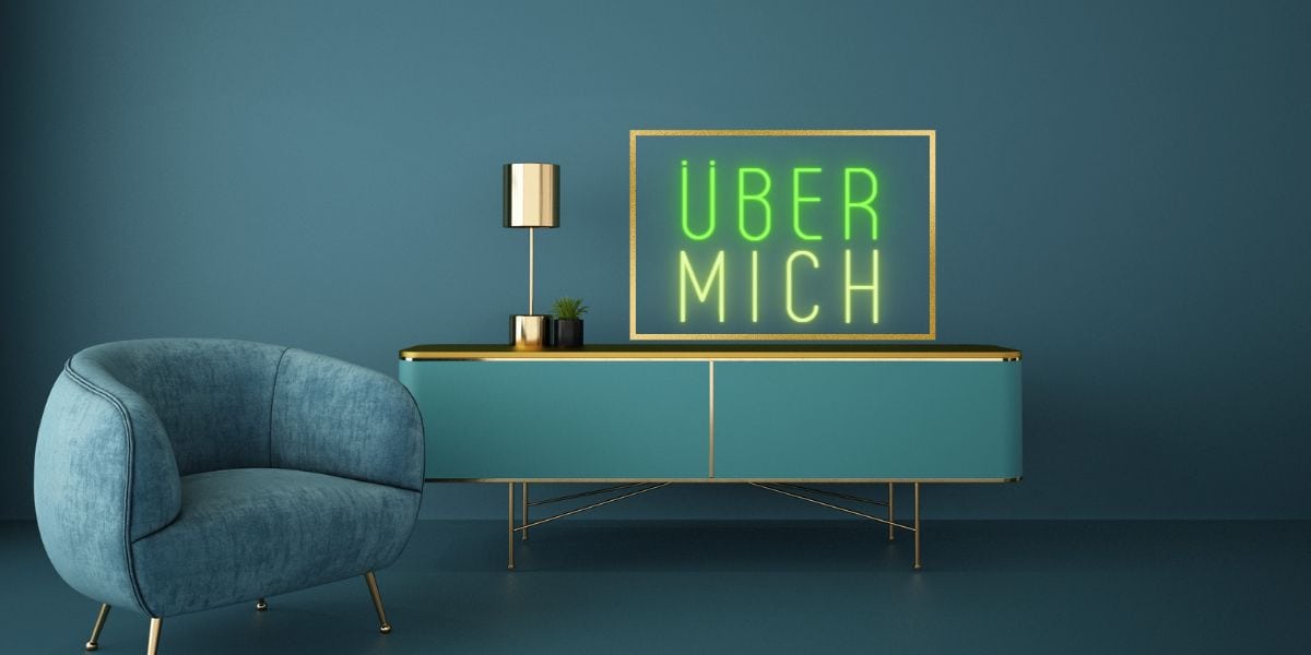 Wichtige Elemente einer Über-mich-Seite, hier: dunkelgrüner Samtsessel, grünes Sideboard mit Goldelementen, goldene Lampe, Bilderrahmen, Schriftzug Über mich, grüne Wände und Teppich