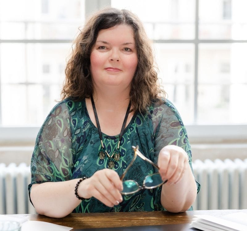 Blog-Expertin Maria Squarra, in grünem Chiffonoberteil am Schreibtisch sitzend, mit Brille in den Händen