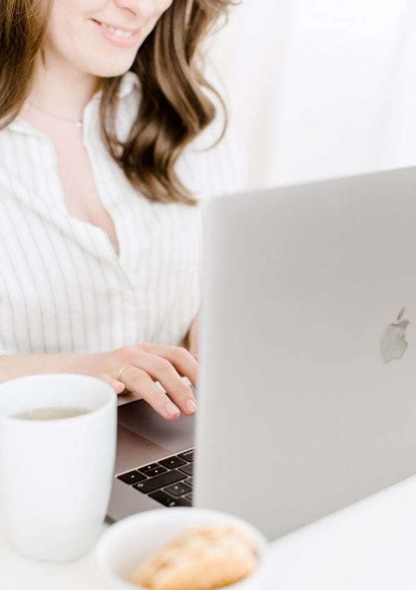 Blog Lounge, hier Frau mit langen dunklen Haaren, weiße Bluse, schreibt am silberfarbenem Laptop, weiße Kaffeetasse, Brioche