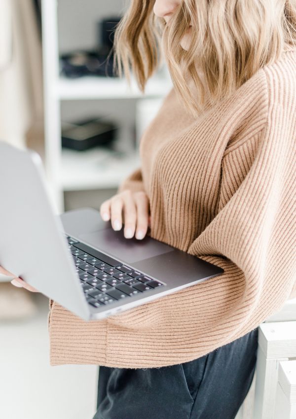 Blog Review, hier Frau im hellbraunen Pullover, stehend, mit offenem Laptop in den Händen