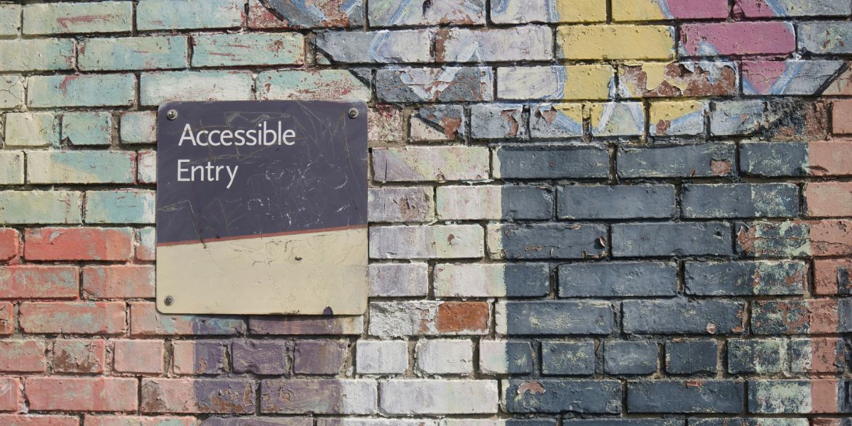 Das Bild "Digitale Barrierefreiheit" zeigt eine alte Ziegelsteinwand, die mit unterschiedlichen Farben bemalt wurde. Ein Schild zeigt die Aufschrift: Accessible Entry.