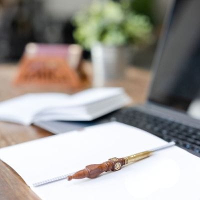 Strategie-Post abonnieren Strategie-Post abonnieren: aufgeschlagenes Bullet Journal mit leeren Seiten, darauf ein Kugelschreiber aus gedrechseltem Holz und Messingspitze, dahinter Laptop in dunkelgrau, kleines geöffnetes Heft, Briefständer aus Sheesham-Holz und kleine Grünpflanze in Zinktopf