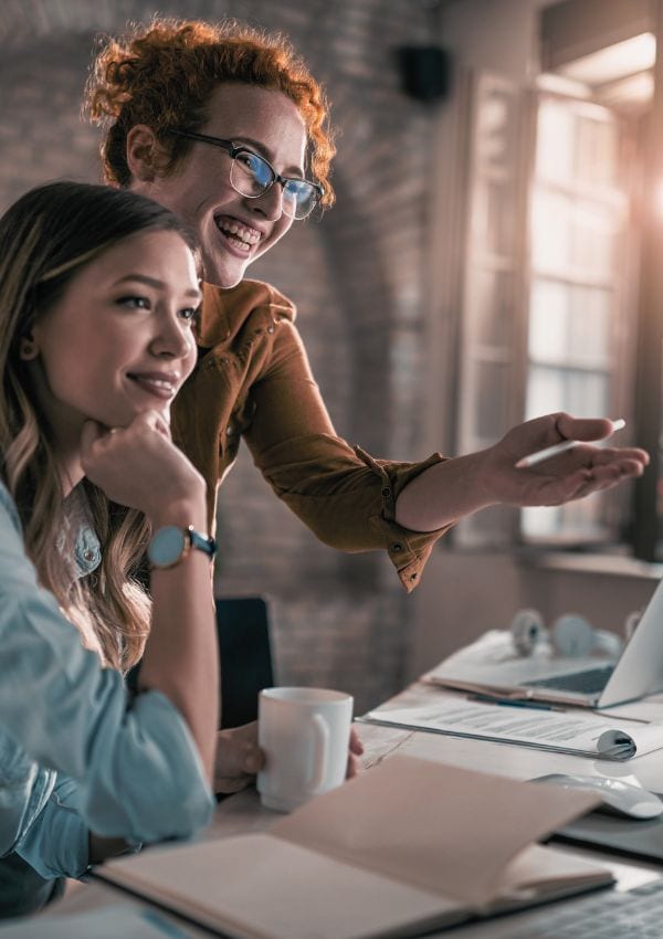 Gemeinsam bloggen in der Blog-Lounge, hier zwei Frauen an einem Schreibtisch mit Notizbuch, Unterlagen, Kaffeebecher, eine Rothaarige stehend und auf einen Laptop weisend, eine Langhaarige sitzend, beide lächeln und schauen auf einen Bildschirm.