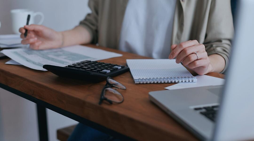 Wechsel zur Kleinunternehmerregelung, hier weibliche Person in weißem T-Shirt und offener schilffarbener Bluse, sitzend an schmalem Holztisch mit Laptop, Taschenrechner, Brille, Kaffeetasse und Unterlagen