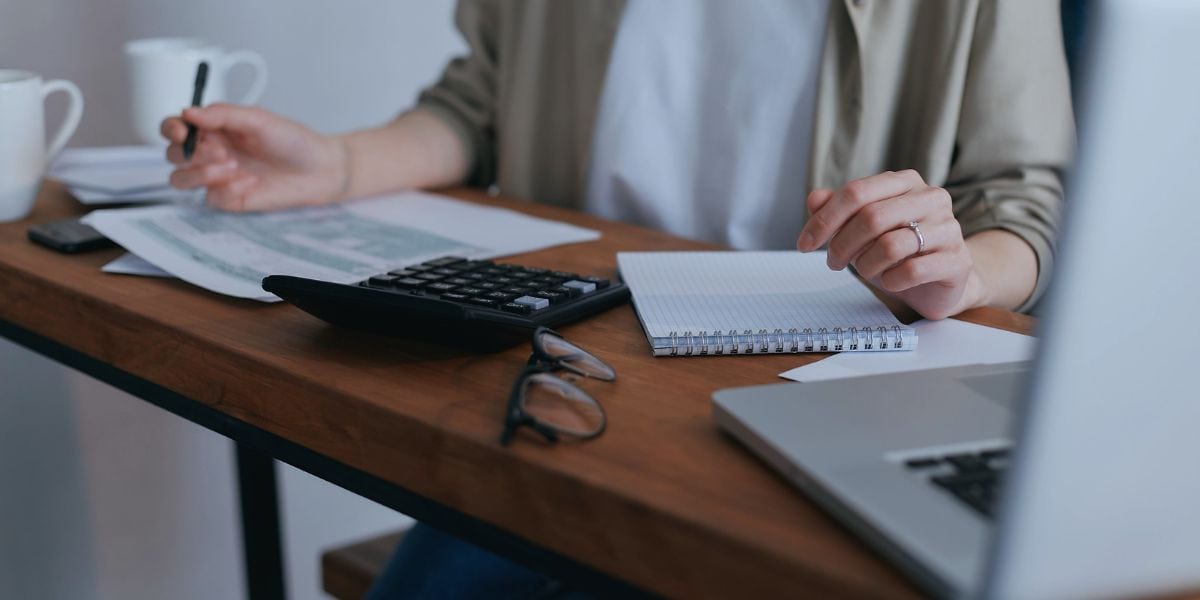 Wechsel zur Kleinunternehmerregelung, hier weibliche Person in weißem T-Shirt und offener schilffarbener Bluse, sitzend an schmalem Holztisch mit Laptop, Taschenrechner, Brille, Kaffeetasse und Unterlagen